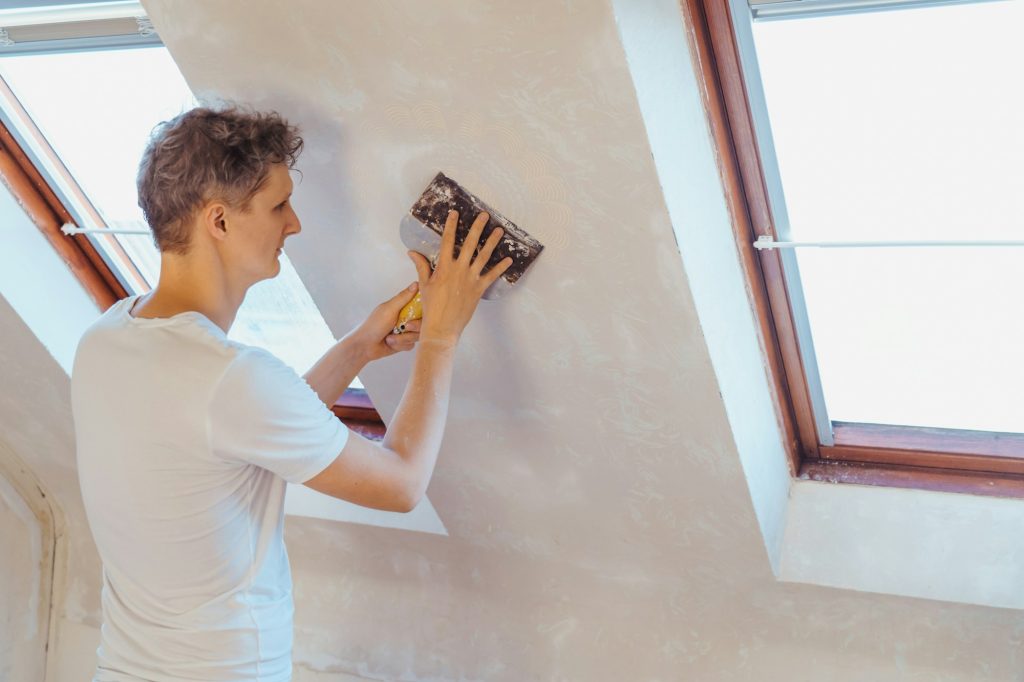 A young man scratching with a spatula and removing old wallpaper on a wall of a room. Do it yourself