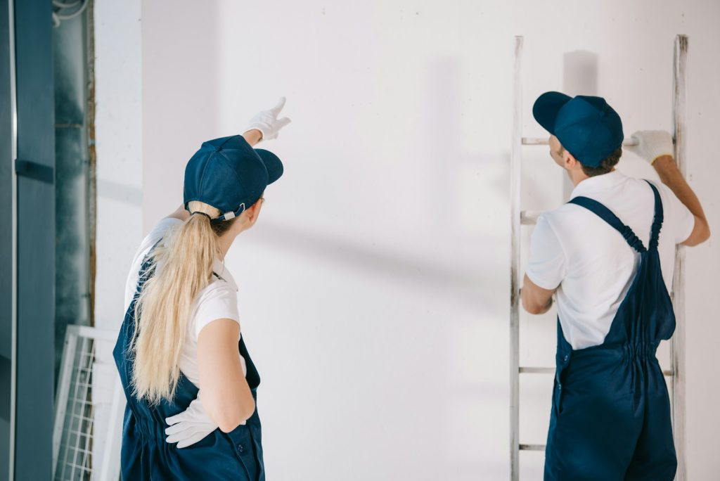 young painter in uniform pointing with finger at white wall while standing near coworker holding